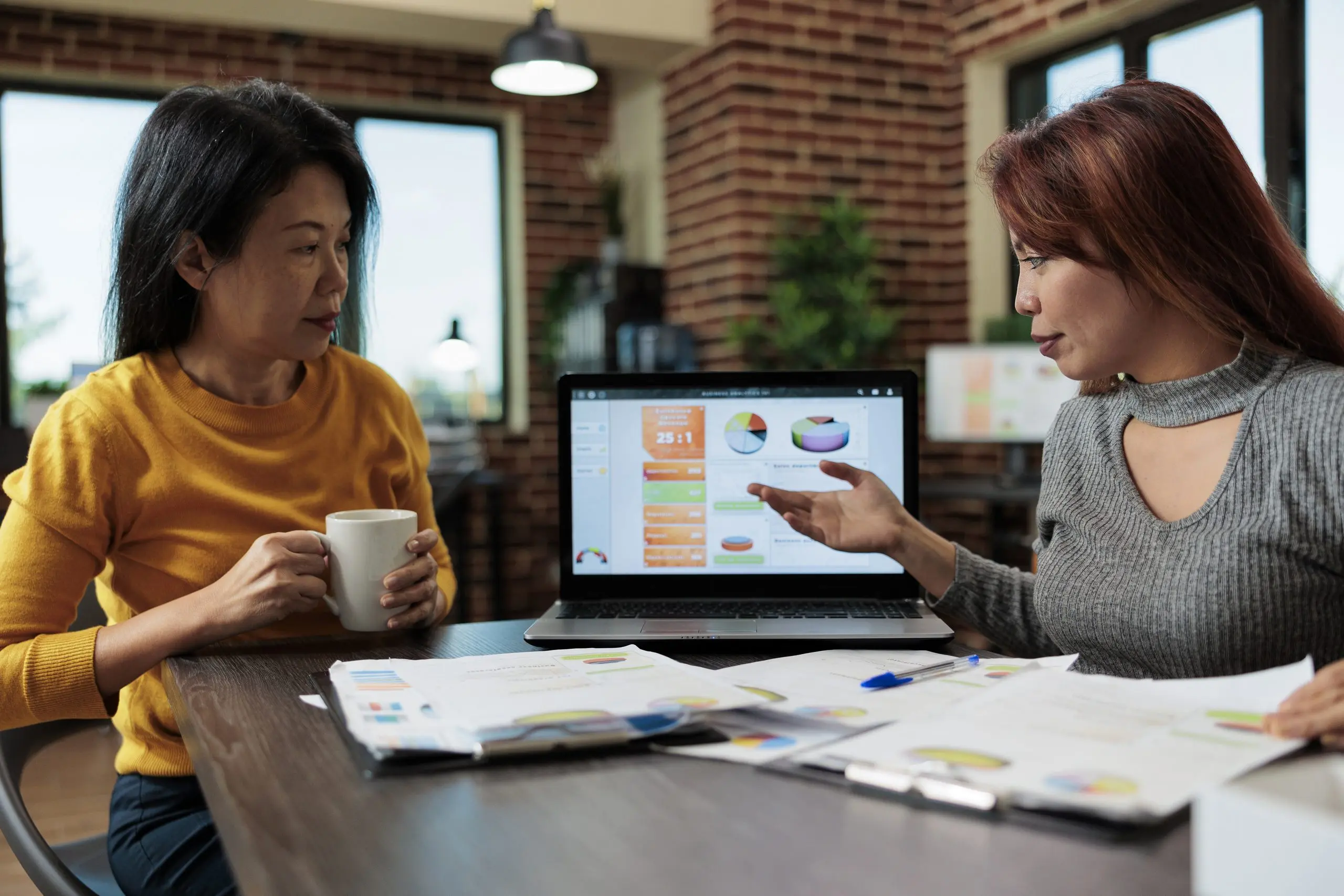 Two women review charts on a laptop and printed reports at a table in a brick-walled office