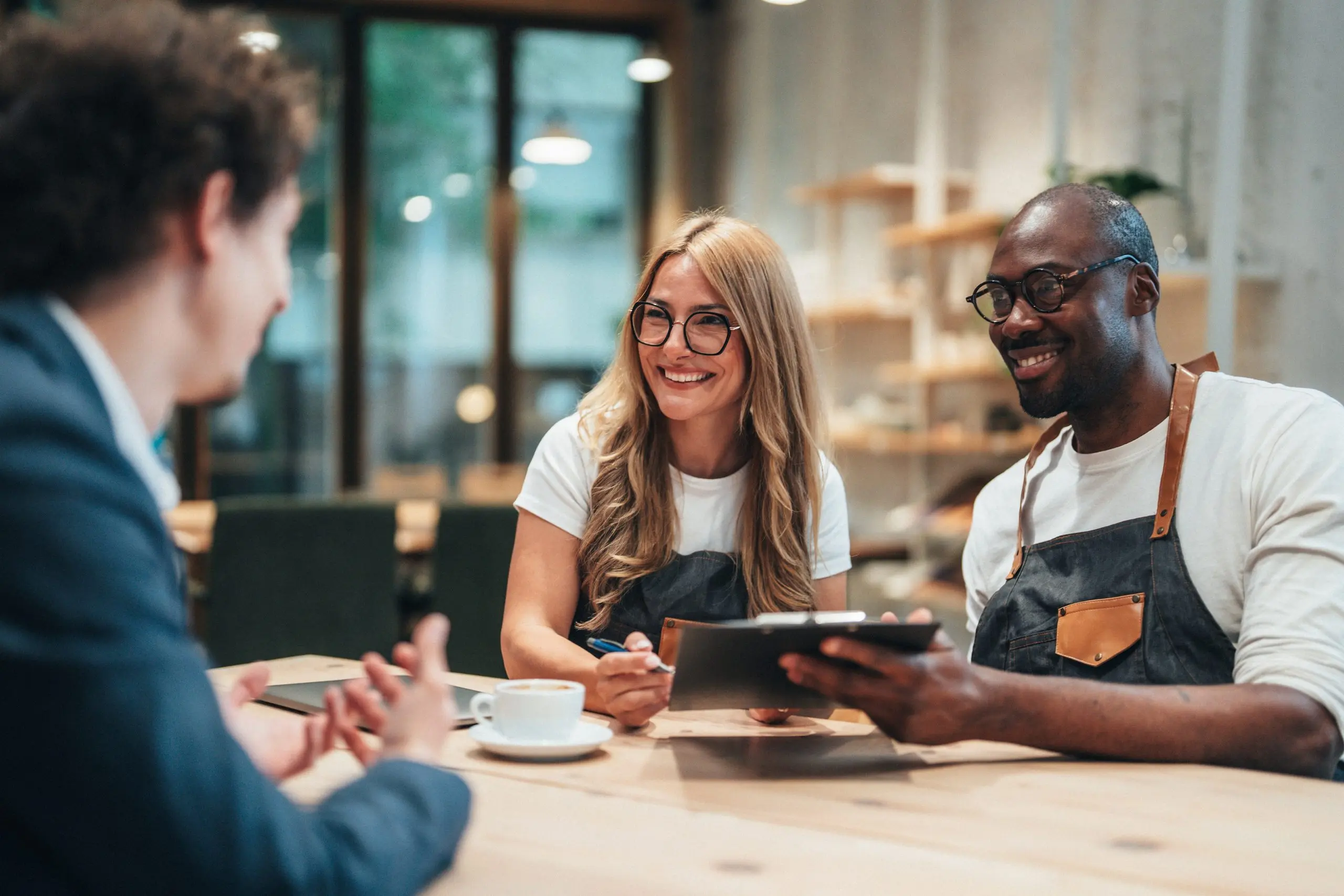 Two small business owners in aprons meet with an adviser at a cafe table with a tablet and coffee