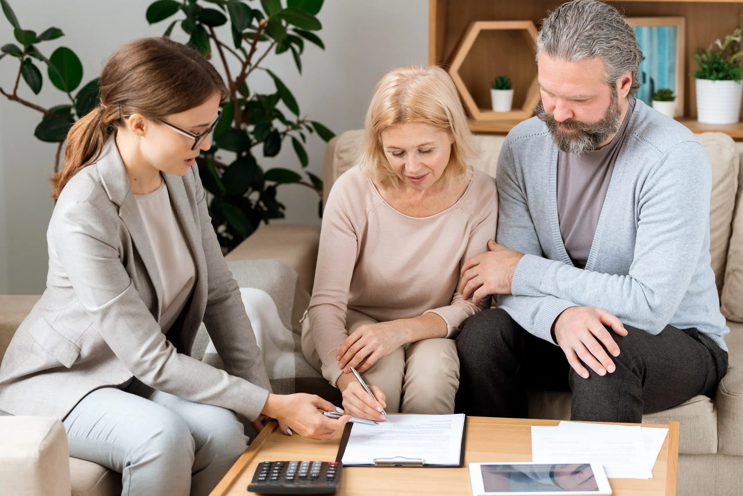 An adviser points to the signature line on a document as a woman signs and a man looks on