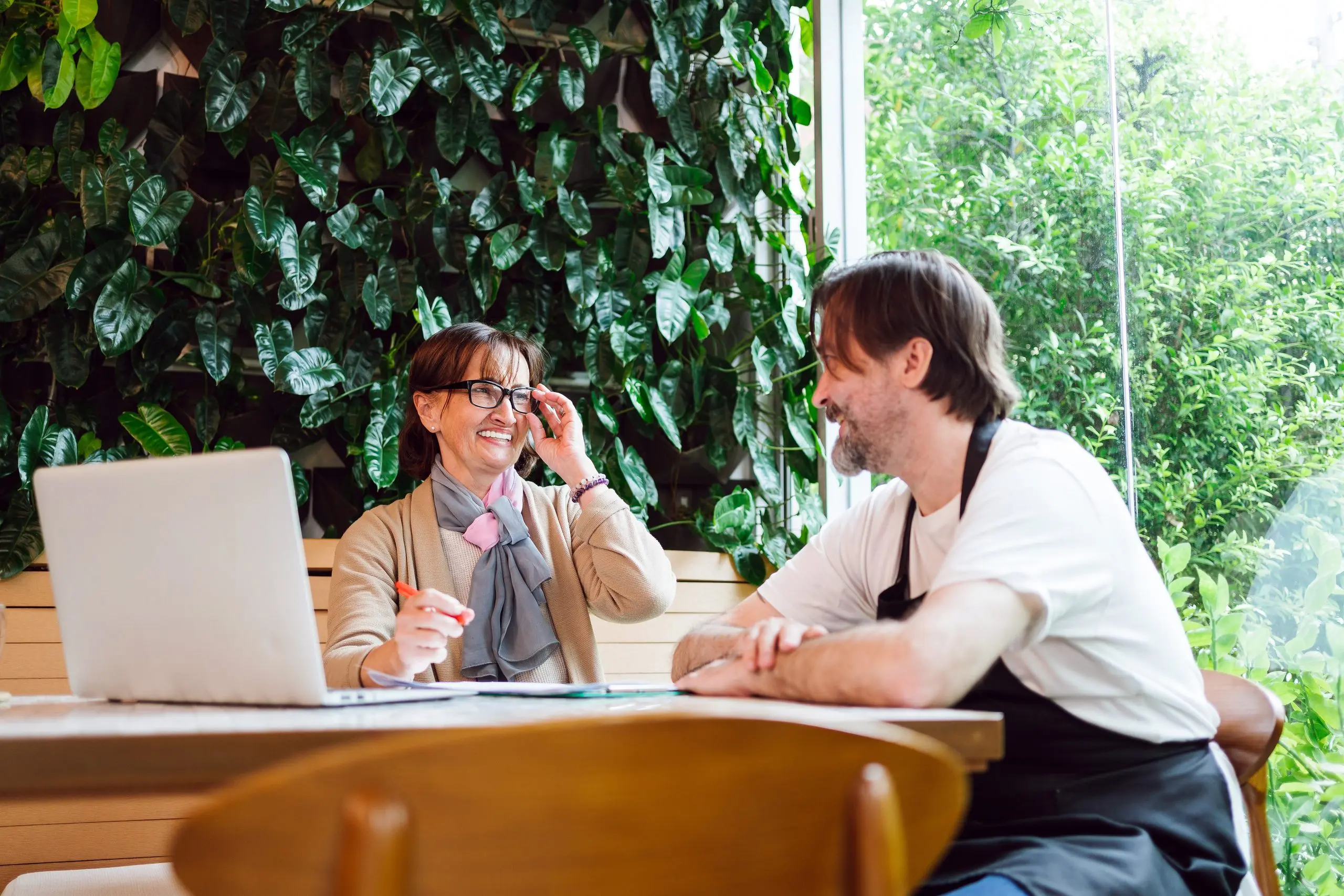 A smiling cafe owner in an apron talks with a woman adviser at a laptop in a plant-filled cafe