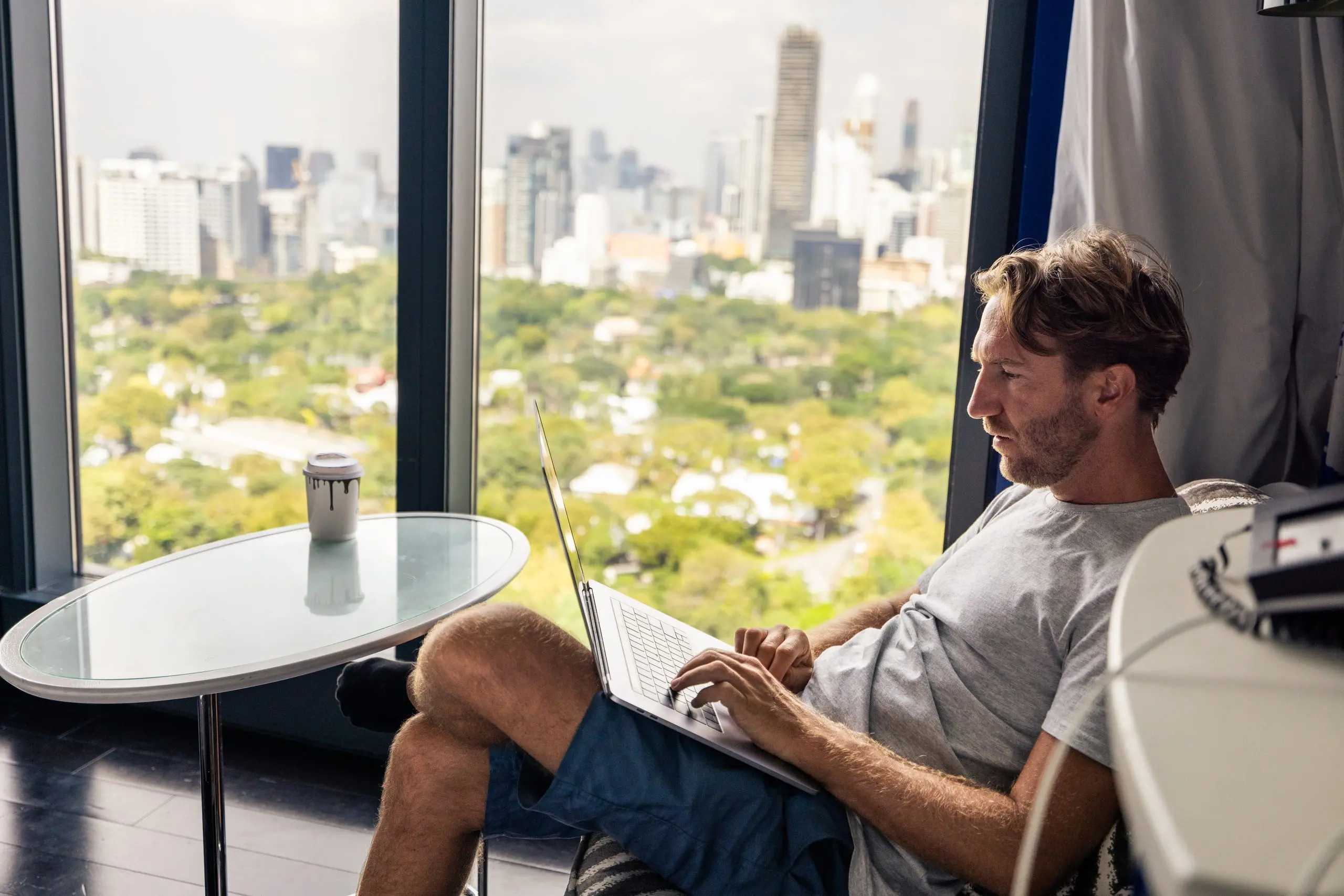 A man works on a laptop in a hotel room with a city skyline outside the window