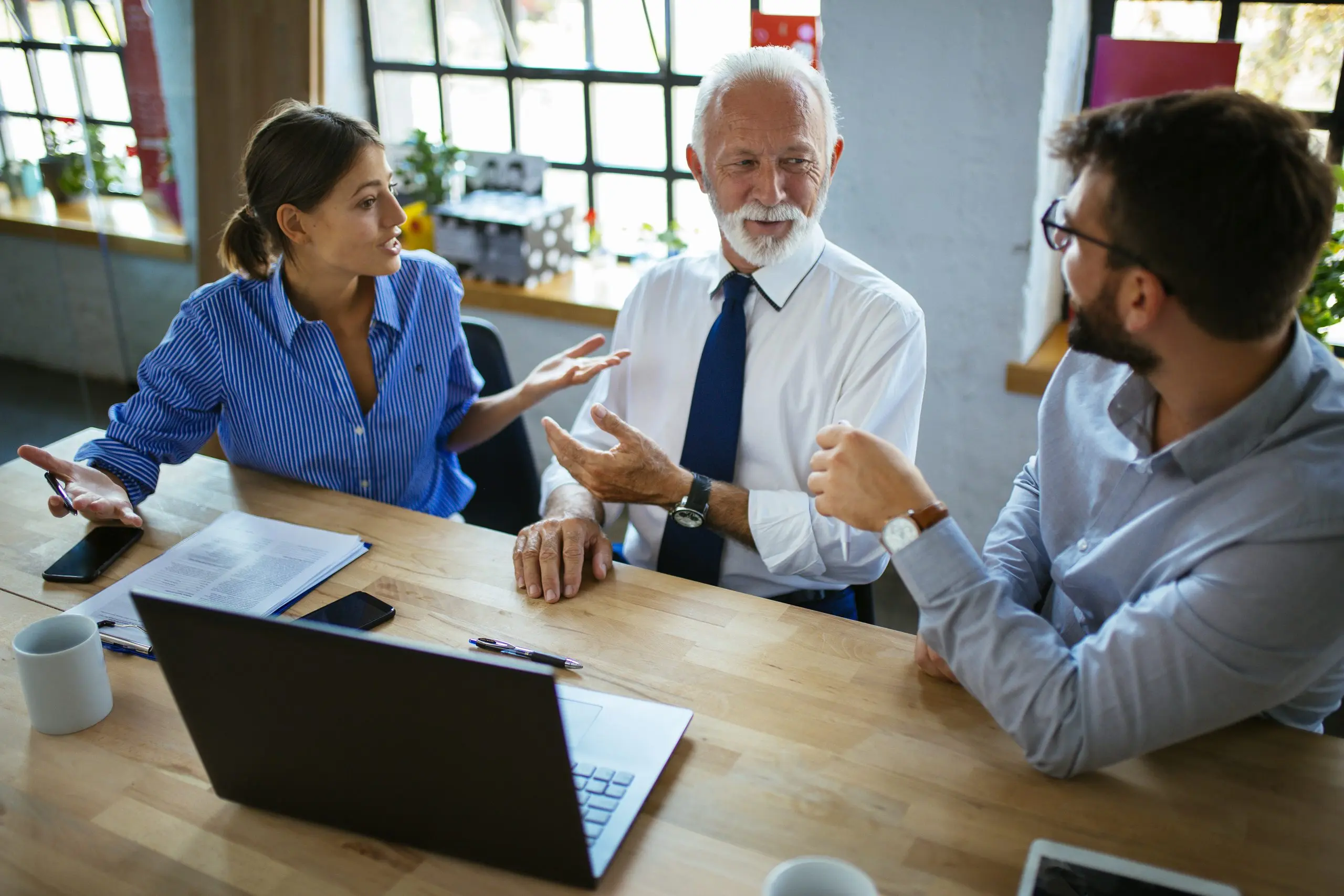 Three colleagues in discussion around a wooden table with a laptop, documents and a phone