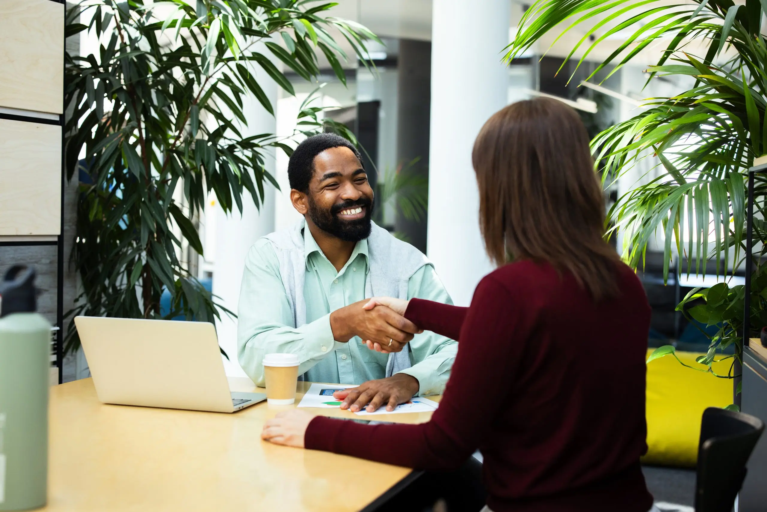 A smiling man shakes hands with a woman across a desk in a modern office with plants